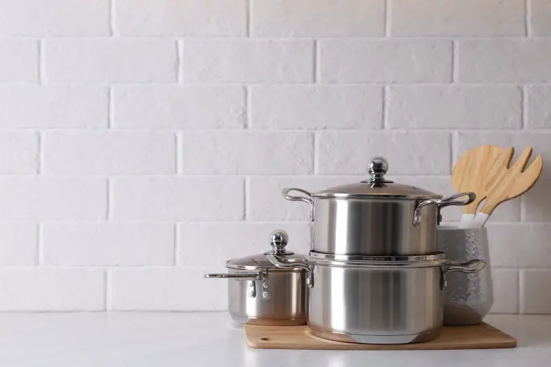 Set of stainless steel cookware and kitchen utensils on table near white brick wall