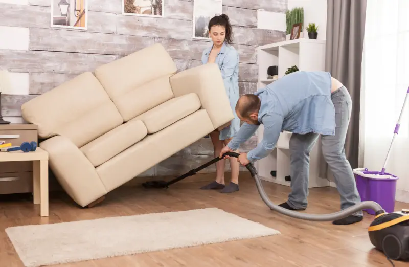 Wife picks up sofa while her husband is cleaning the dust under it with vacuum
